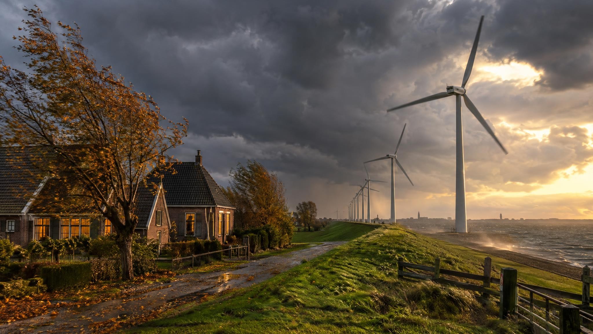 Windturbines draaien bij harde wind langs een dijk, met Nederlandse huizen en buigende bomen onder een donkere herfstlucht
